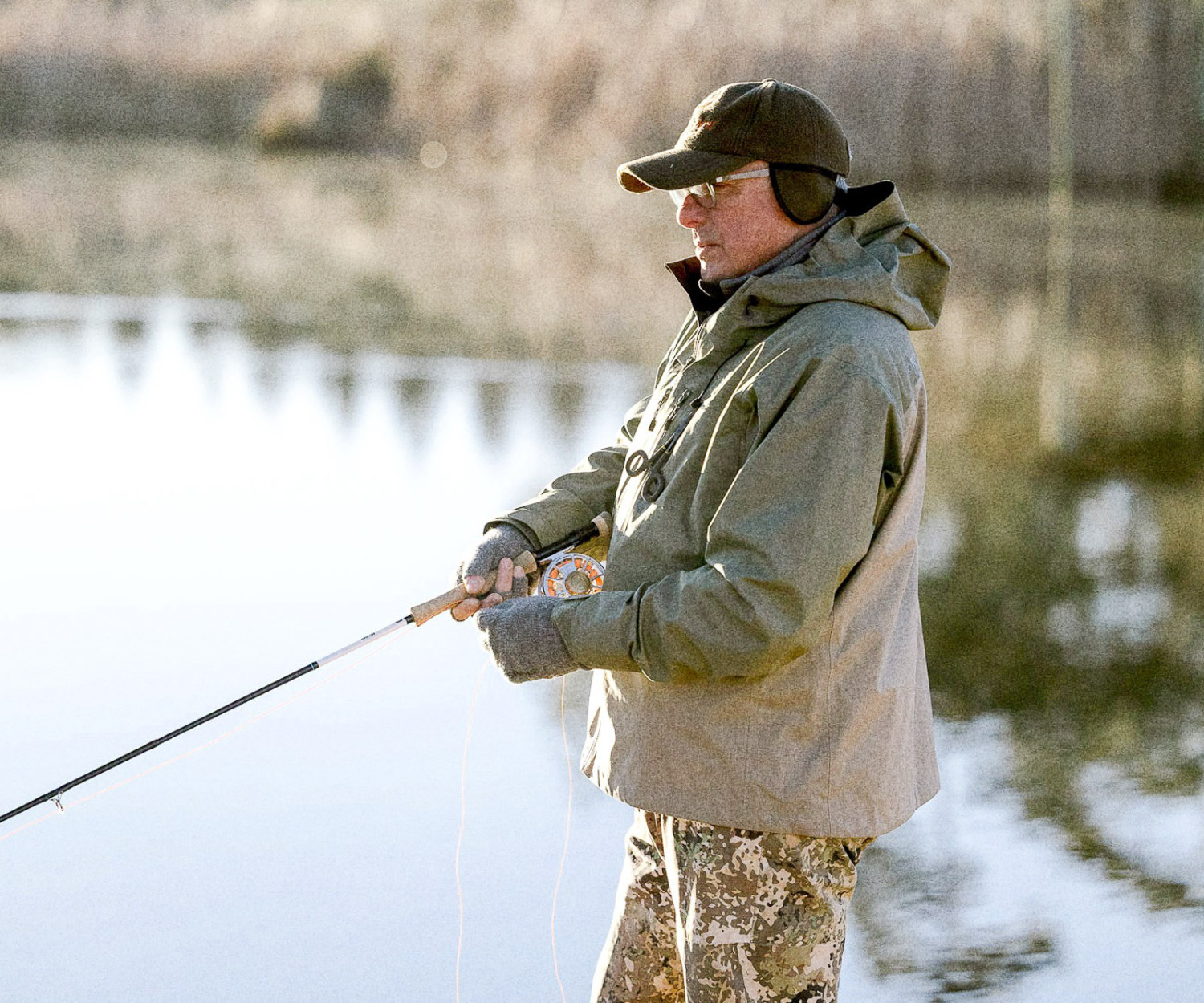 Mavungana fly fishing: Man fly fishing in nature. Angler wearing a jacket, hat, and gloves, holding a fishing rod near water.